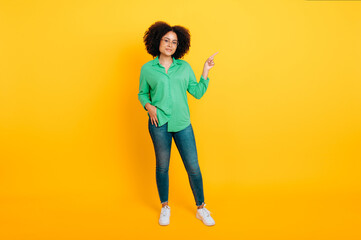 Fototapeta na wymiar Full length photo of positive pretty brazilian or hispanic curly woman, points with finger to the side at empty space for advertise or announcement, smile at camera, isolated yellow background