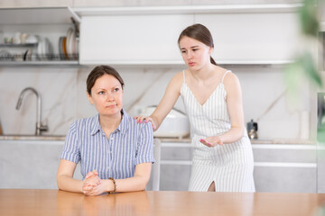 Offended mother sitting at the kitchen table, her young daughter standing behind and trying to talk...
