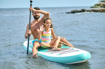 Father and daughter surfing on sup board and having funny time, family summer vacations.