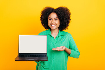 Obraz na płótnie Canvas Joyful brazilian or african american curly woman, in green shirt, holding an open laptop in hand with white blank mock up screen and points hand at it, smile at camera, isolated yellow background