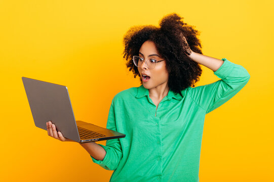 Joyful surprised brazilian or african american curly haired woman, in a green shirt, holding an open laptop in hand, looks in amazement at it, holding hand on head, stand on isolated yellow background