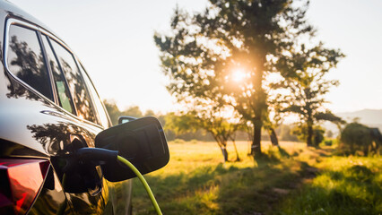 Black electric car with the power cable supply plugged in, in the blurred background visible green area and trees. Eco friendly and renewable energy concept.