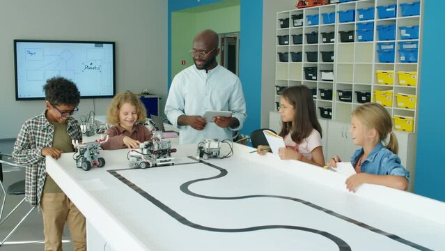 Full shot of group of diverse casually dressed preteen kids and African American teacher standing around table, watching and testing electric robot models at after school technology club