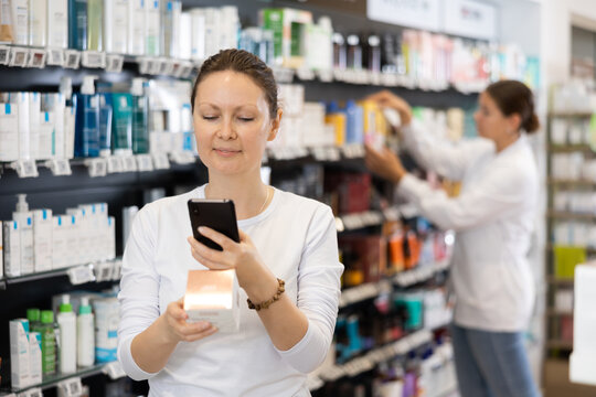 Adult woman in casual clothes looking for cosmetic product from photo on internet on smartphone in pharmacy..