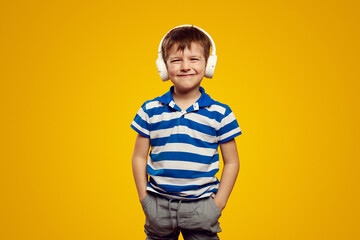 Playful boy in headphones listening to music and smiling while holding hands in pockets on yellow background.