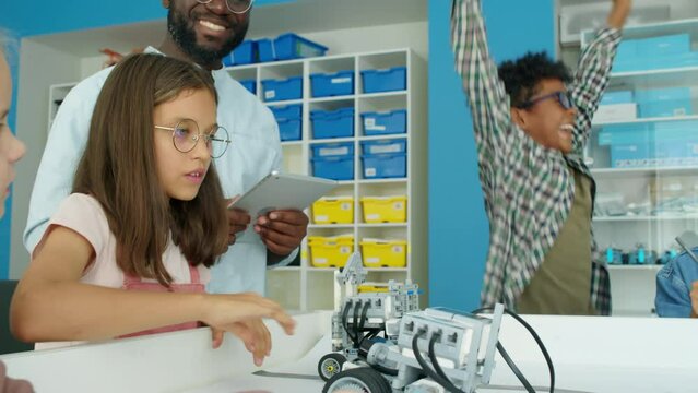 Medium shot of group of young diverse children and black male teacher playing with programmable electric robots at after school tech club, and ecstatic boy jumping with joy at completing task