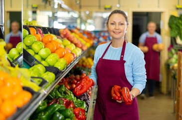 Positive middle-aged woman market assistant arranging organic bell pepper in grocery shop