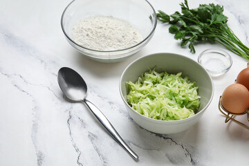 Bowls with grated zucchini, flour and eggs for preparing fritters on white marble background