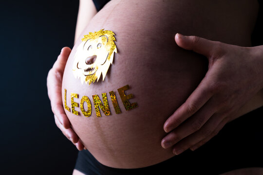 Baby bump shot with glittering lion sticker and child name from the side of pregnant woman at home. Last month of pregnancy - week 36. Black background.
