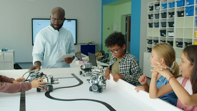 Medium shot of group of preteen diverse schoolchildren and black male IT teacher with tablet computer watching programmable electric robot models wheel around on table at youth robotics club