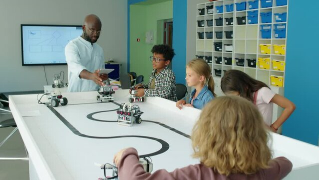 Medium full shot of four young diverse children playing with electric toy robots at after school technology club, and African American male teacher with tablet computer giving directions