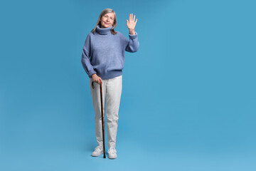 Senior woman with walking cane waving on light blue background © New Africa