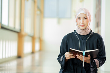 The image of an Asian Muslim woman in the Islamic religion in hijab in white color. reading the Quran, Koran Staying in a beautiful mosque, Arabic word translation: The Holy Al Quran.