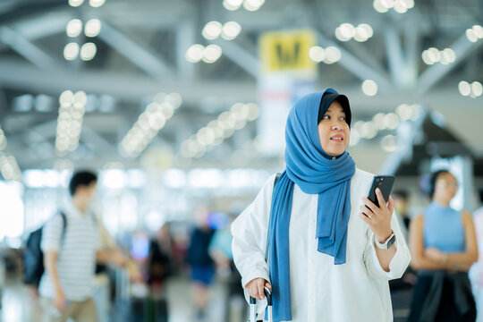 An Asian Muslim Wearing A Blue Hijab Is Preparing For A Vacation And She Is At The Airport. She Is Using Her Mobile Phone To Contact Her Friends And Muslim Travelers.