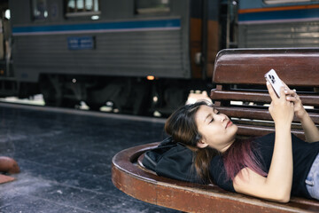 Young women traveler lying down while holding a smartphone waiting for train at the train station