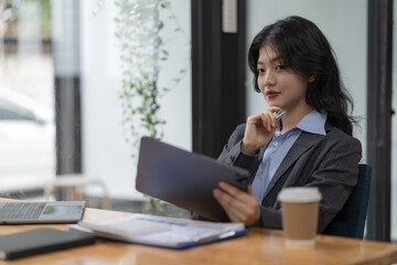 Attractive business Asian woman using a digital tablet while sitting at a workplace desk in an office.