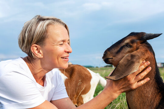 Laughing young woman in a white tank top with a goat in a field. Love and tenderness for animals and nature. Close-up.
