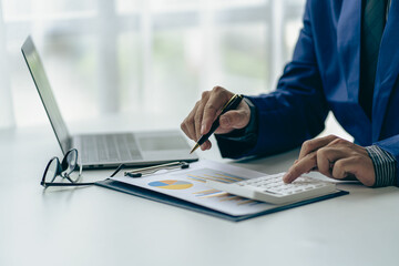 Businessman holding pen pointing to market graph and working on laptop computer working using calculator for mathematics on wooden table at office finance concept close up pictures