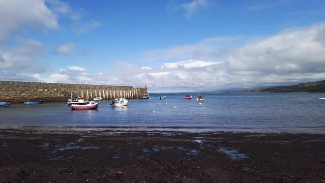 Trefor harbour at ground level panning left to right with view of harbour wall and boats at low tide with backdrop of blue skies on a summers day across the Irish Sea on the Llyn Peninsula in Wales