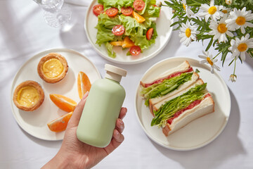 Healthy summer salad vegetarian meal concept, fresh salad with tomato, sandwiches, orange and cake on white background. Female hand is holding green bottle unbranded, product mockup for advertising