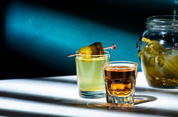 Pickleback, alcoholic cocktail drink from cucumber pickle and whiskey in small shot glasses. Hard light and shadow pattern, green background
