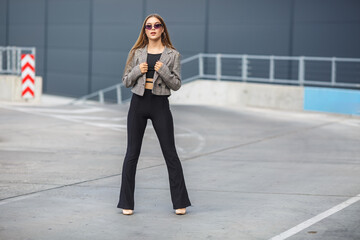 beautiful young girl with sunglasses in jacket posing near a modern office building with mirrored walls in a parking lot with road signs