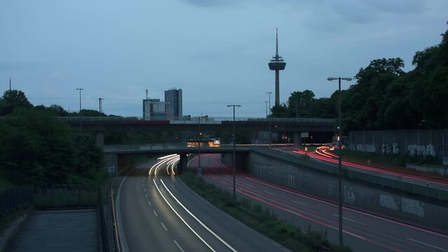 Traffic Flow and Cologne Skyline. Witness bustling cars with light strokes on a highway, a prominent TV tower, moving clouds, and Cologne's panoramic skyline in the evening. Energy and dynamism. Z002