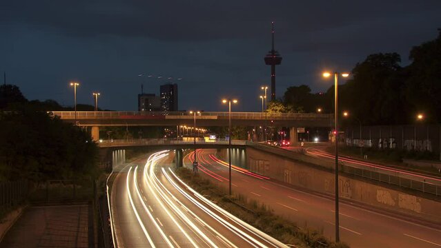 Traffic Flow and Cologne Skyline. Witness bustling cars with light strokes on a highway, a prominent TV tower, moving clouds, and Cologne's panoramic skyline in the evening. Energy and dynamism. Z003