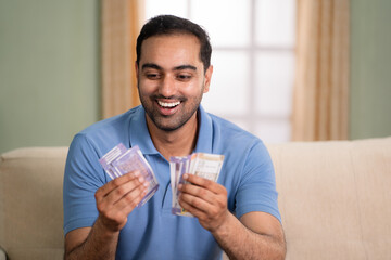 Happy excited indian man counting money or currency notes while sitting on sofa at home - concept...