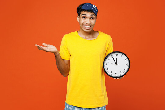 Sad sleepy young man he wear pyjamas jam sleep eye mask rest relax at home hold clock spread hand say oops being late isolated on plain orange background studio portrait. Good mood night nap concept.