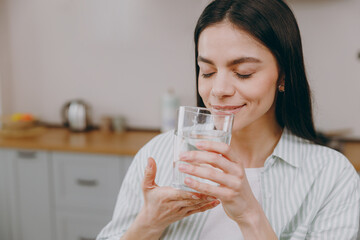 Close up young smiling relaxed satisfied cheerful happy housewife latin woman wears casual clothes striped shirt drink water sit on table in light kitchen at home alone Lifestyle cooking food concept