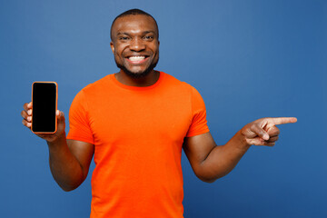 Obraz na płótnie Canvas Young man of African American ethnicity he wear orange t-shirt hold in hand use mobile cell phone with blank screen workspace area point aside isolated on plain dark royal navy blue background studio