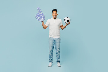 Full body young man fan wear basic t-shirt foam 1 fan glove finger up cheer up support football sport team hold in hand soccer ball watch tv live stream isolated on plain pastel blue color background. - obrazy, fototapety, plakaty
