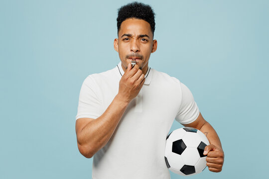 Young strict referee man fan wear basic t-shirt whistle look camera cheer up support football sport team hold in hand soccer ball watch tv live stream isolated on plain pastel blue color background.