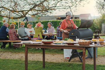 Senior Woman Barbecuing at Countryside Gathering - A senior woman prepares food at a barbecue as...