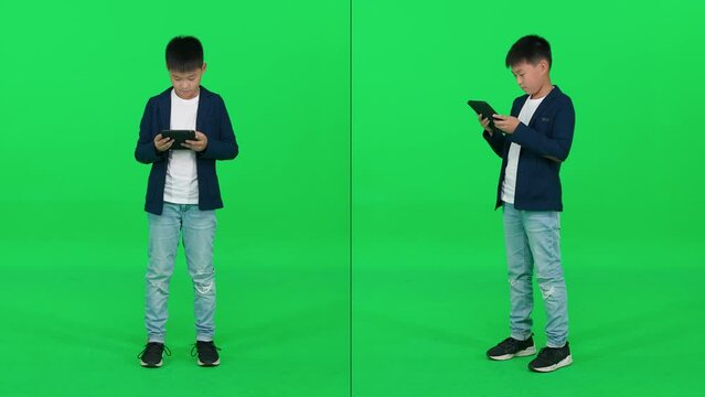 Focused asian schoolboy in a suit standing on a green background and using a screen tablet, reads an e-book, video collage template on a chromakey.