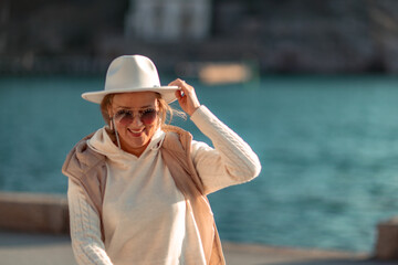 Happy blonde woman in a white suit and hat posing at the camera against the backdrop of the sea