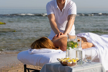 Woman having relax in tropical massage spa on the beach near the sea on massage table. Professional masseur provides back and neck spa procedures.