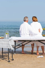 Obraz premium Family – man and woman sitting on massage table and looking at the sea after having relax procedures in tropical massage spa on the beach.
