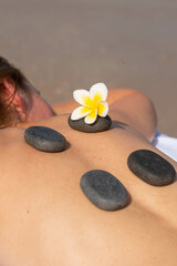 Obraz na płótnie Canvas Massage hot stones on woman’s back with tropical flower. Woman having relax in tropical massage spa on the beach near the sea on massage table.