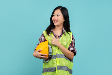 Portrait of a beautiful Asian woman engineer wearing hardhat and vest pointing a finger in an empty space.