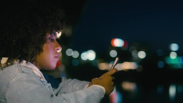 Black woman standing on the bridge at night and browsing her mobile phone. Shot with RED helium camera in 8K. 