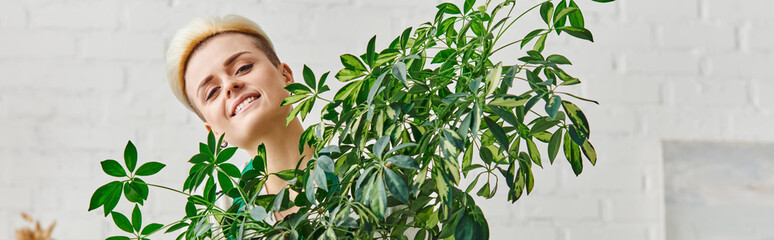 portrait of young woman with happy smile and trendy hairstyle looking at camera near green houseplant in living room, indoor gardening, sustainable home decor and green living concept, banner