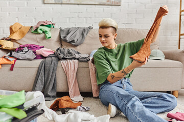 Obraz premium young woman sitting on floor with suede boot near clothes and couch at home, sorting and reducing wardrobe, trendy hairstyle, tattoo, sustainable living and mindful consumerism concept