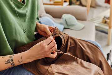 cropped view of young tattooed woman buttoning leather pants while sitting on couch near wardrobe items during decluttering process, sustainable living and mindful consumerism concept