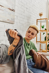 Fototapeta na wymiar tattooed woman with trendy hairstyle sorting clothes and reducing wardrobe items on couch in living room near rack with green plants at home, sustainable living and mindful consumerism concept