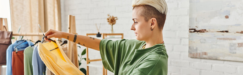 clothing sorting, side view of young tattooed woman with trendy hairstyle standing with yellow jumper near rack with garments on hangers, sustainable living and mindful consumerism concept, banner