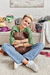 Obraz premium wardrobe items sorting, happy tattooed woman sitting on floor with leather bag and looking at camera, trendy hairstyle, casual clothes, sustainable living and mindful consumerism concept
