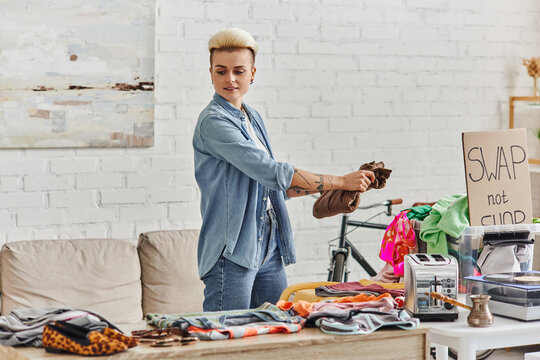 stylish and tattooed woman holding wardrobe item near second-hand clothes, electric toaster, plastic container, cezve and swap not shop card, sustainable living and circular economy concept