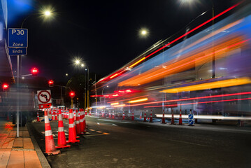 Orange traffic cones lined up on the road. Bus light trails approaching red traffic lights....